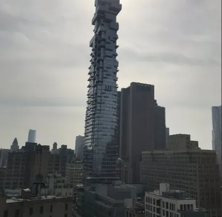 Stacked glass tower rising above lower Manhattan rooftops on a hazy day