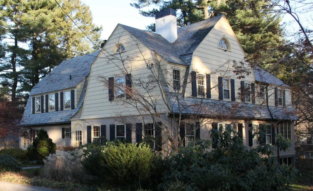 Gambrel-roofed Colonial Revival house with dark shutters behind garden plantings