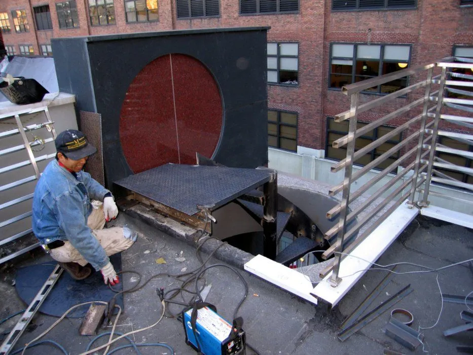 Welder fitting a steel spiral stair beside a round red door on a city rooftop