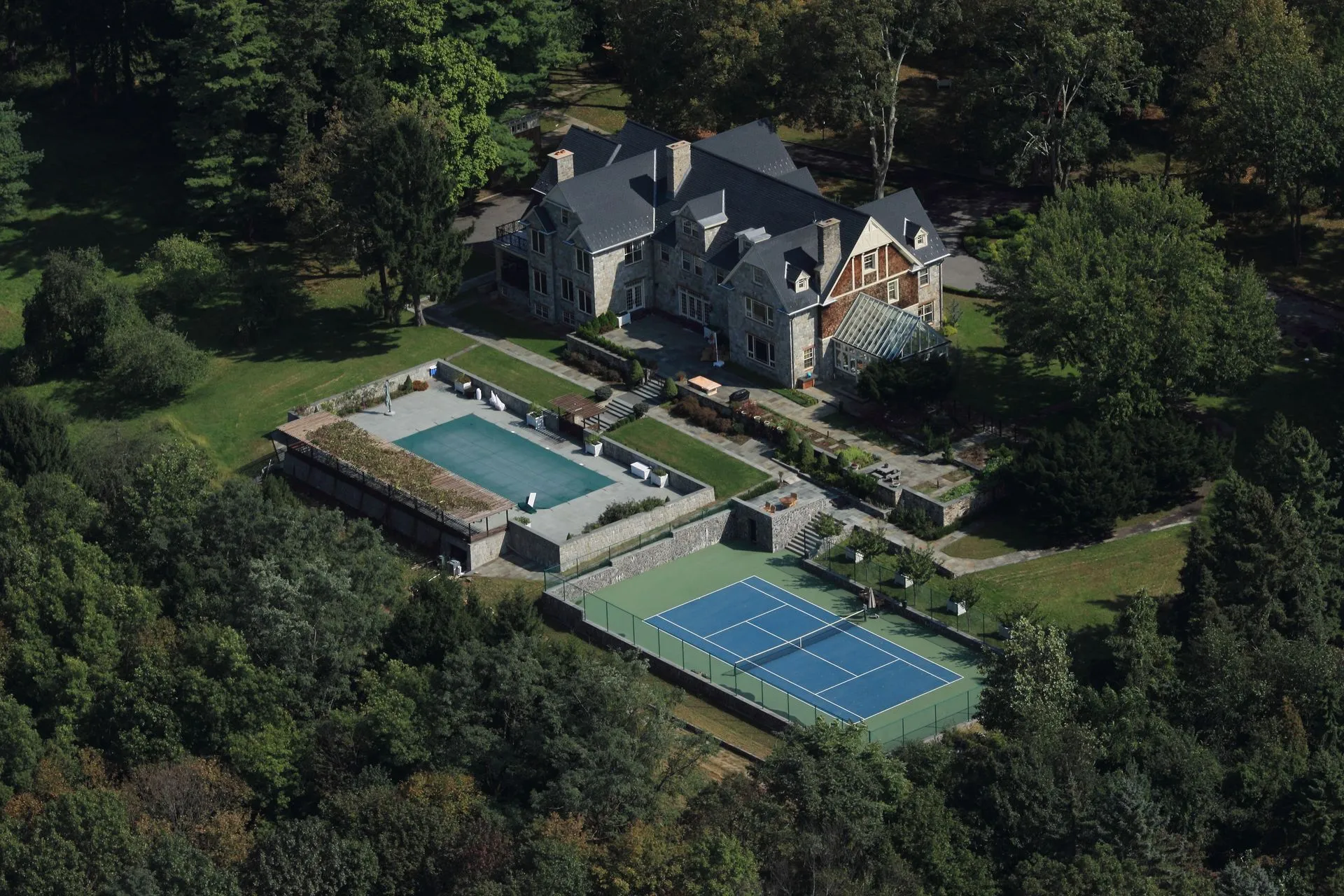 Aerial view of a stone estate house with a terraced pool and tennis court in the woods